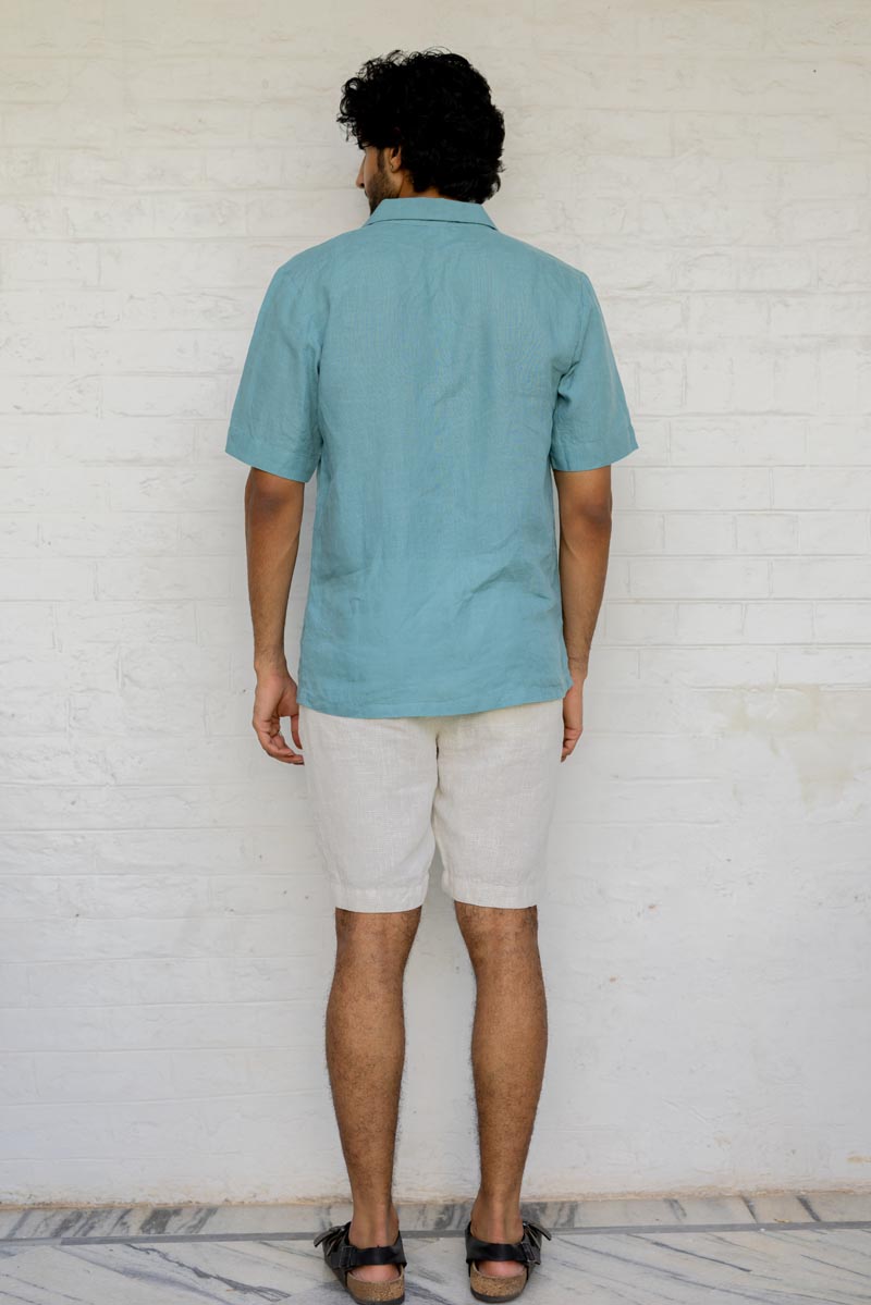 Man wearing a Cuban Collar Turquoise Shirt and beige shorts standing against a white brick wall.