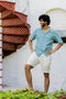 Man wearing a Cuban Collar Turquoise Shirt and white shorts standing in front of a white wall with a red spiral staircase.