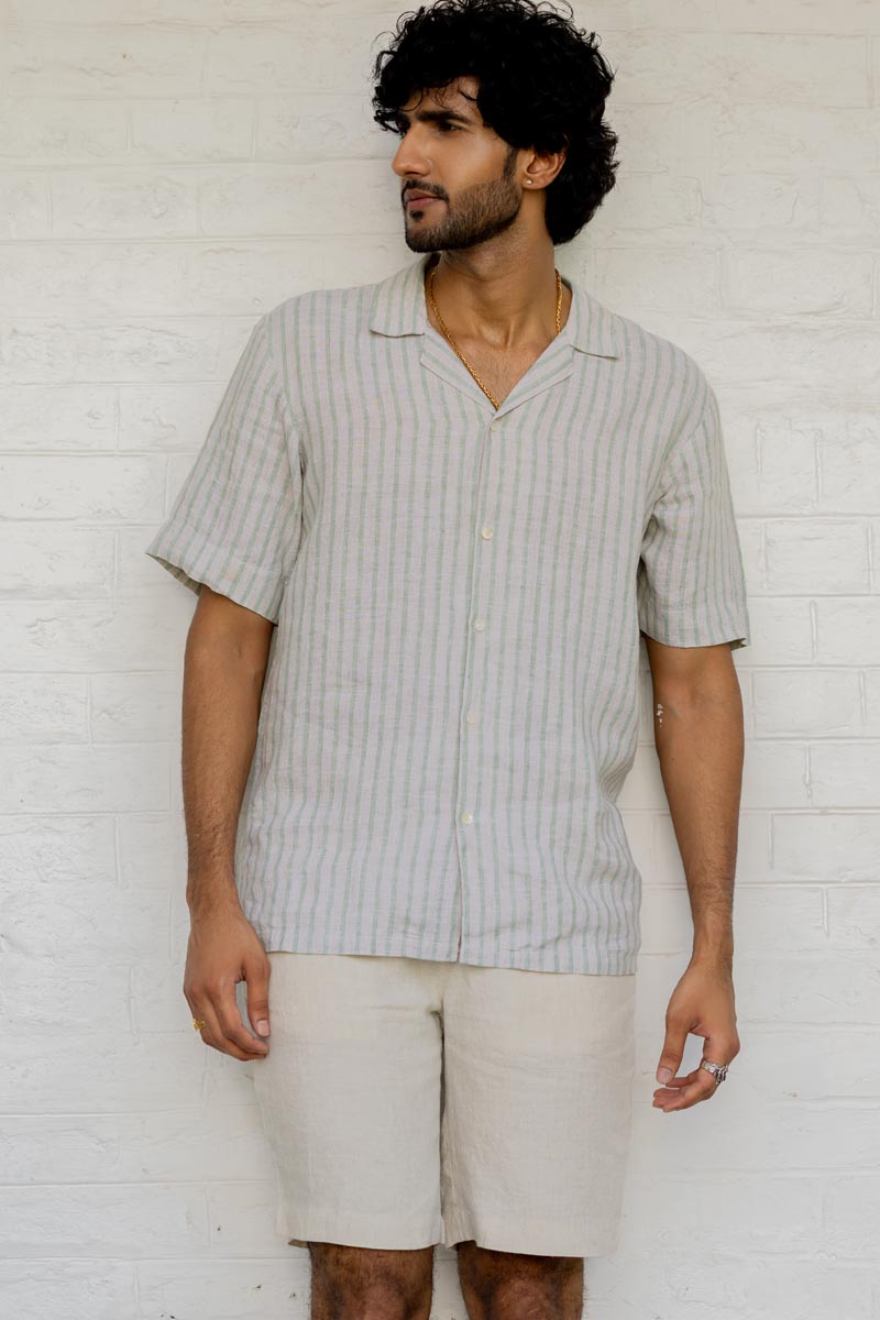Man wearing a Cuban Collar Shirt Sage Pencil Stripe and beige shorts against a white brick wall.