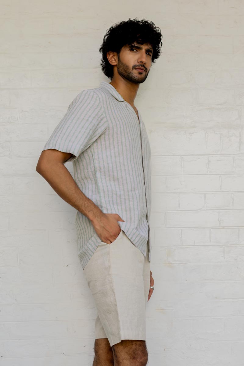 Man wearing a Cuban Collar Shirt Sage Pencil Stripe and shorts against a white brick wall.