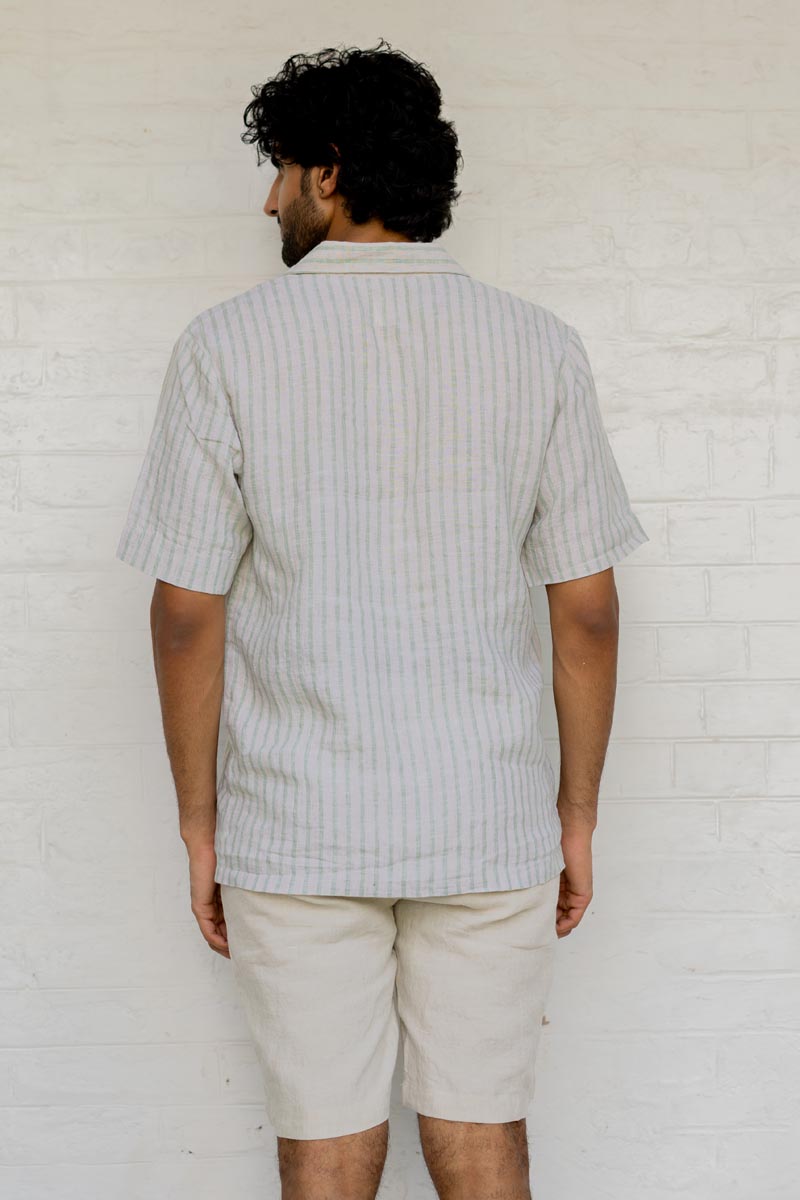 Man wearing a Cuban Collar Shirt Sage Pencil Stripe and beige shorts against a white brick wall.