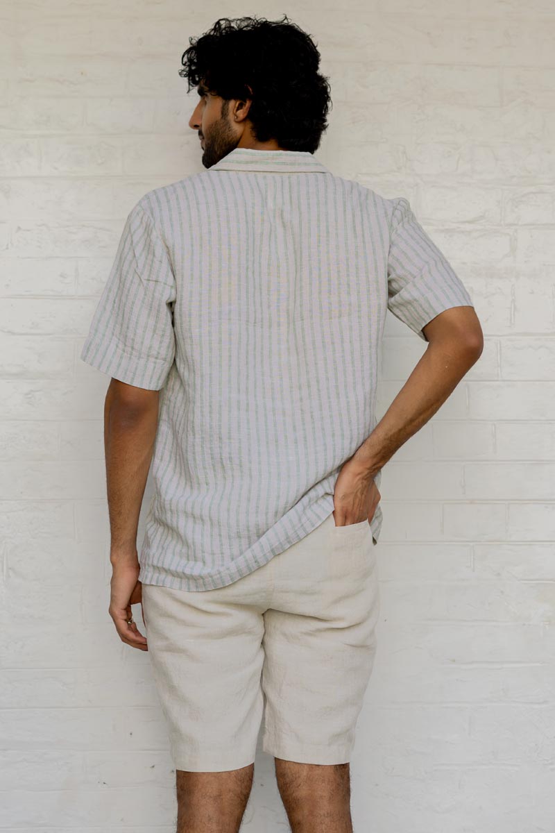 Man wearing a Cuban Collar Shirt Sage Pencil Stripe and beige shorts against a white brick wall.