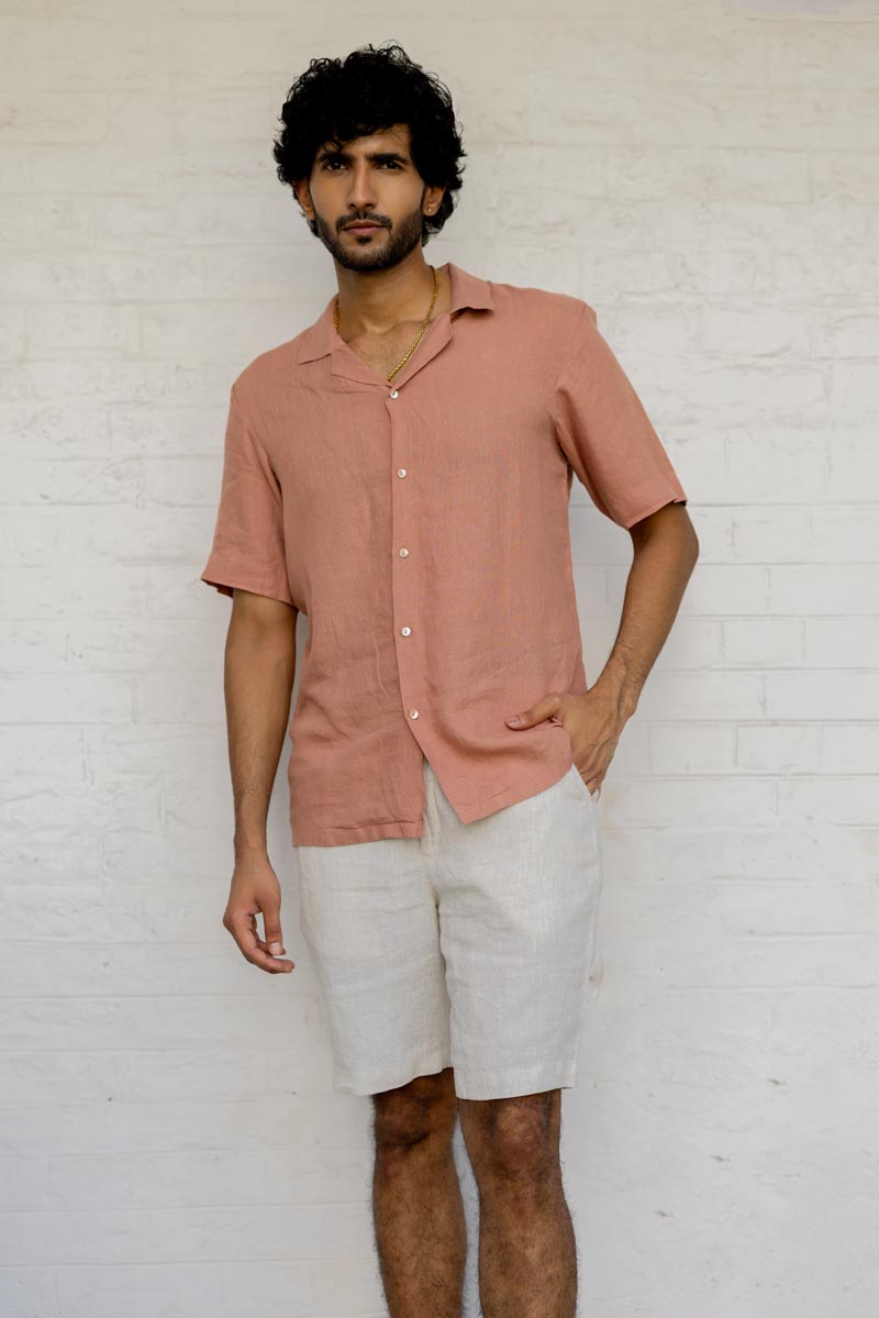 Man wearing a Cuban Collar Apricot Shirt and white shorts against a white brick wall.
