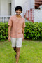 Man Cuban Collar Apricot Shirt and light shorts standing on grass with a building and bushes in the background