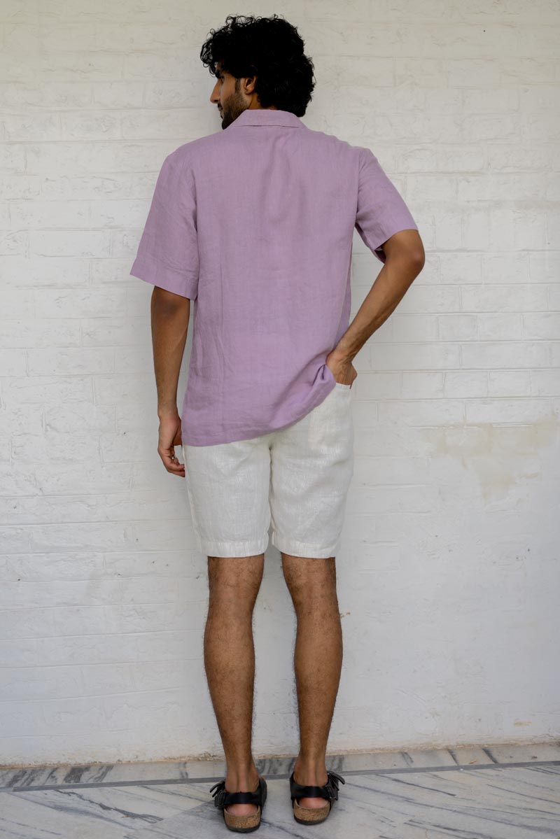 Man wearing a Cuban Collar Light Lilac Shirt and white shorts standing against a white brick wall.