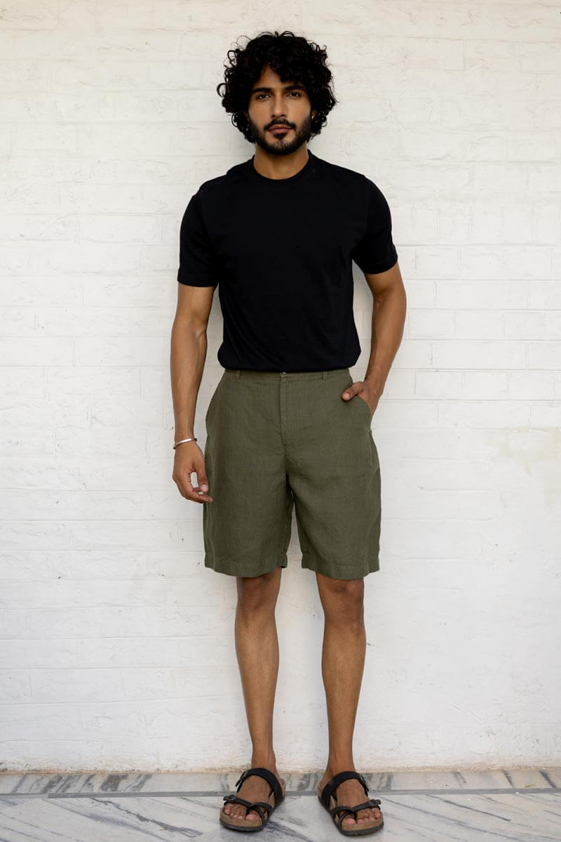Man wearing a black t-shirt and Crocodile Green Shorts standing against a white brick wall.