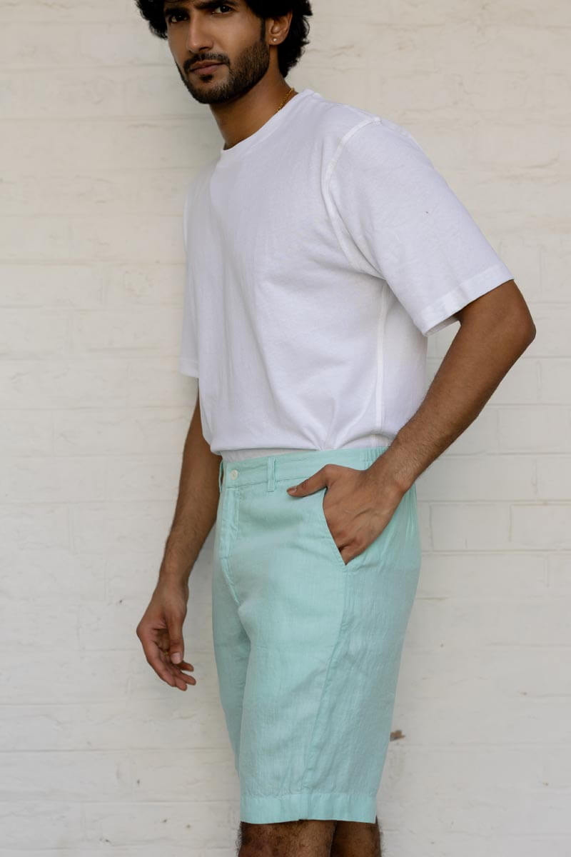 Man wearing a white t-shirt and Celestial Green Shorts against a white brick wall.