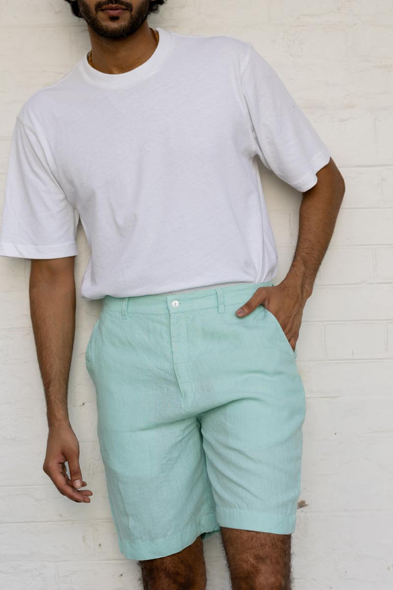 Person wearing a light gray t-shirt and Celestial Green Shorts against a white brick wall.